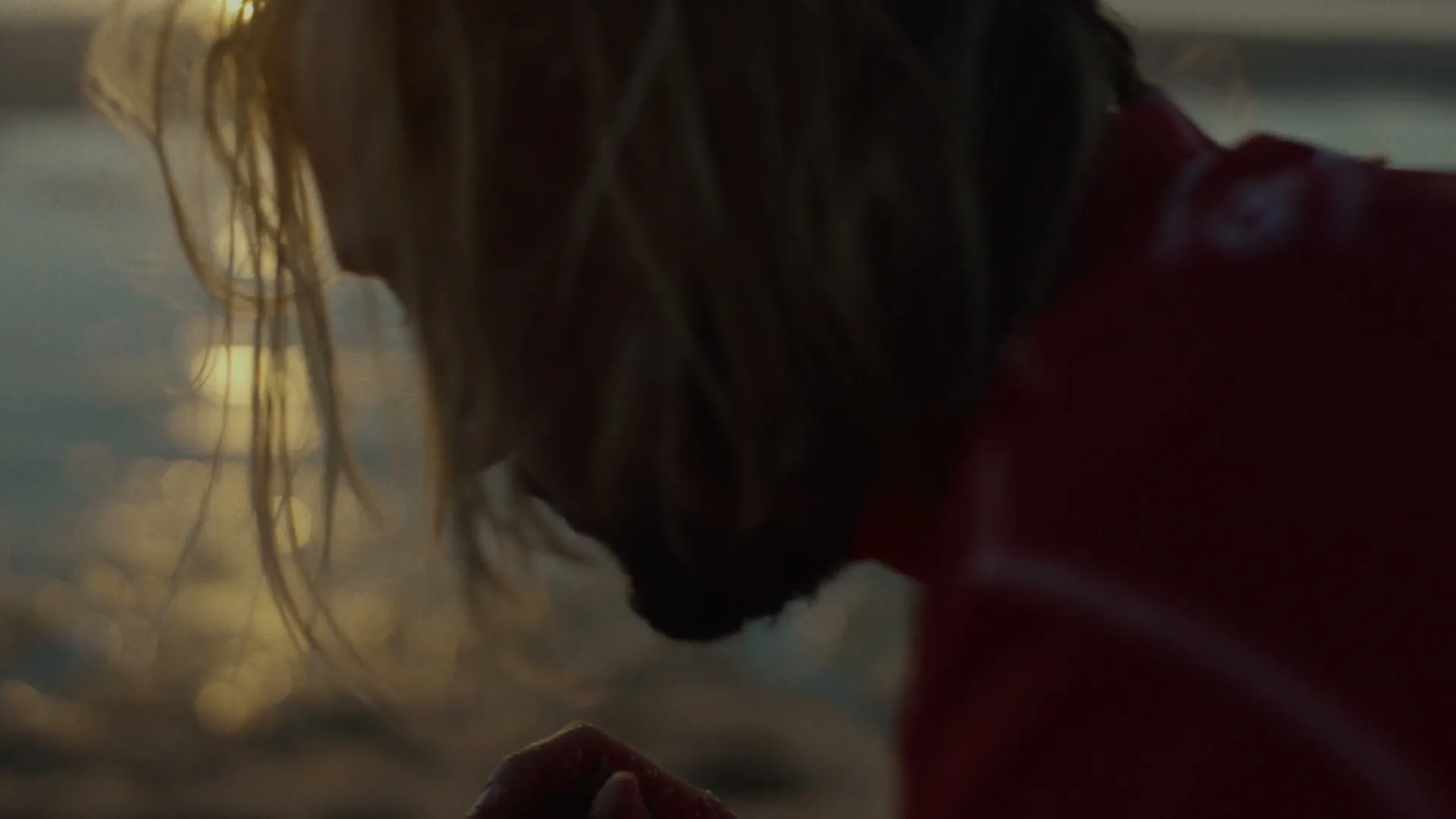 A surfer with wet blonde hair and a red wetsuit bends down on a sandy beach at sunset, with the ocean reflecting the golden light in the background.