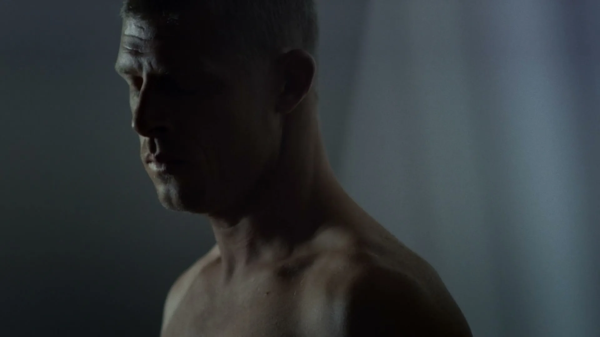 A shirtless man with short grey hair looks down in a dark, moody interior setting.