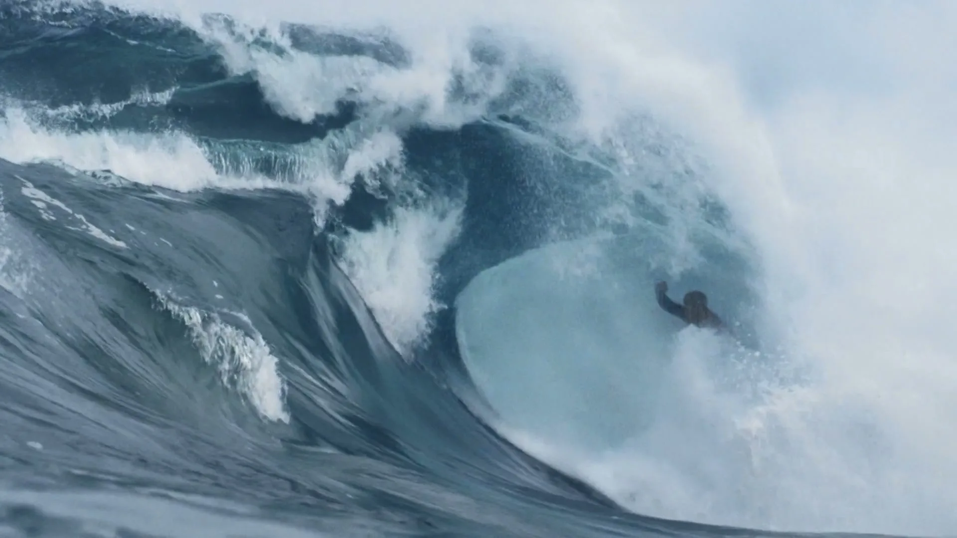 A surfer in a dark wetsuit is engulfed by a massive, breaking blue ocean wave under a cloudy sky.