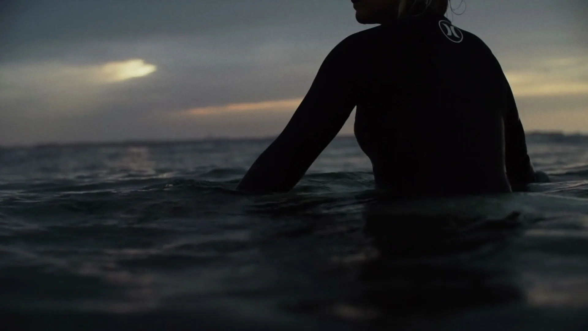 A female surfer in a black wetsuit sits in the ocean, looking out at the waves under a cloudy, dark sky at dusk.