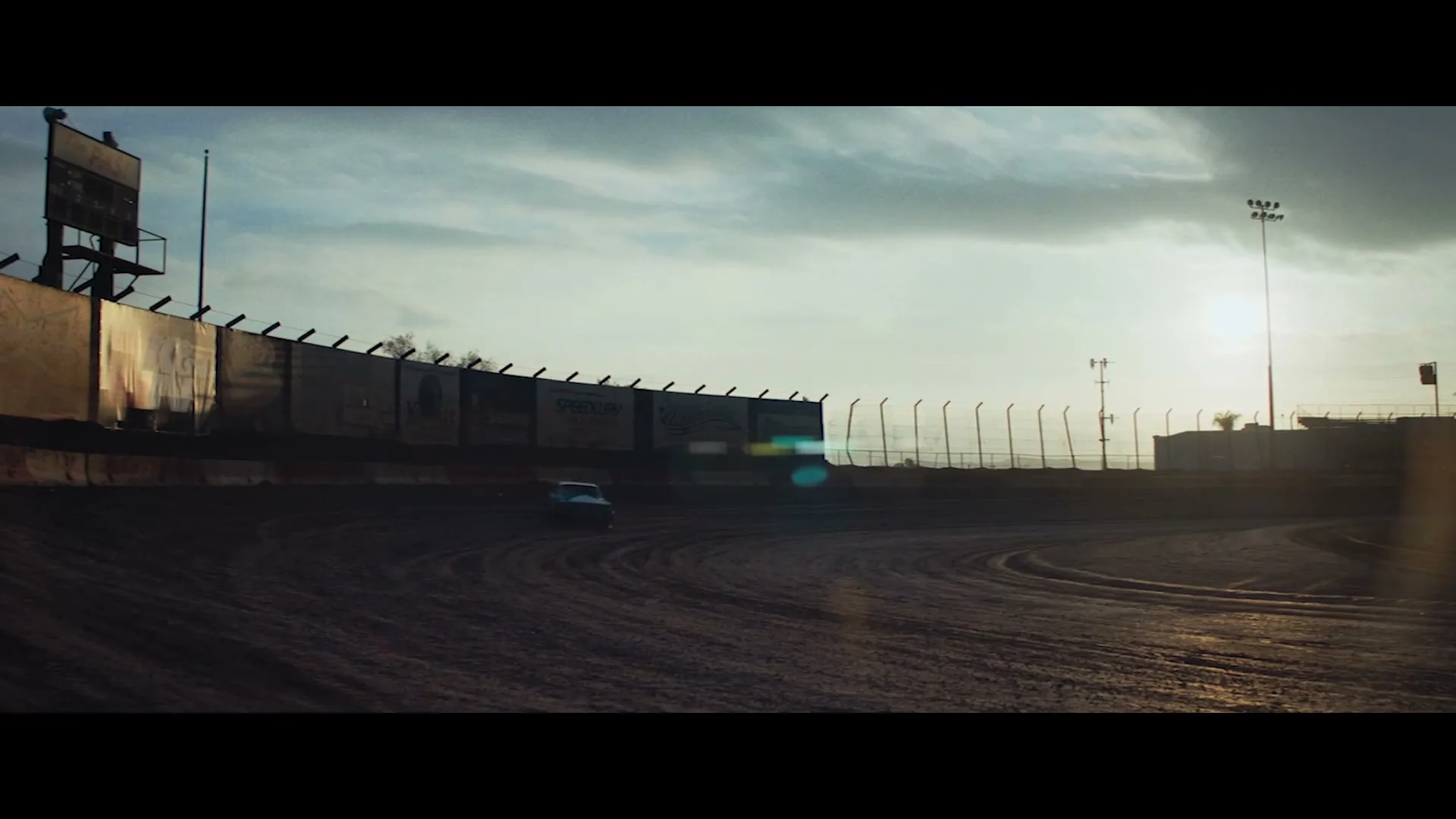 A blue stock car races around a dirt track, kicking up dust as the sun sets in the background.