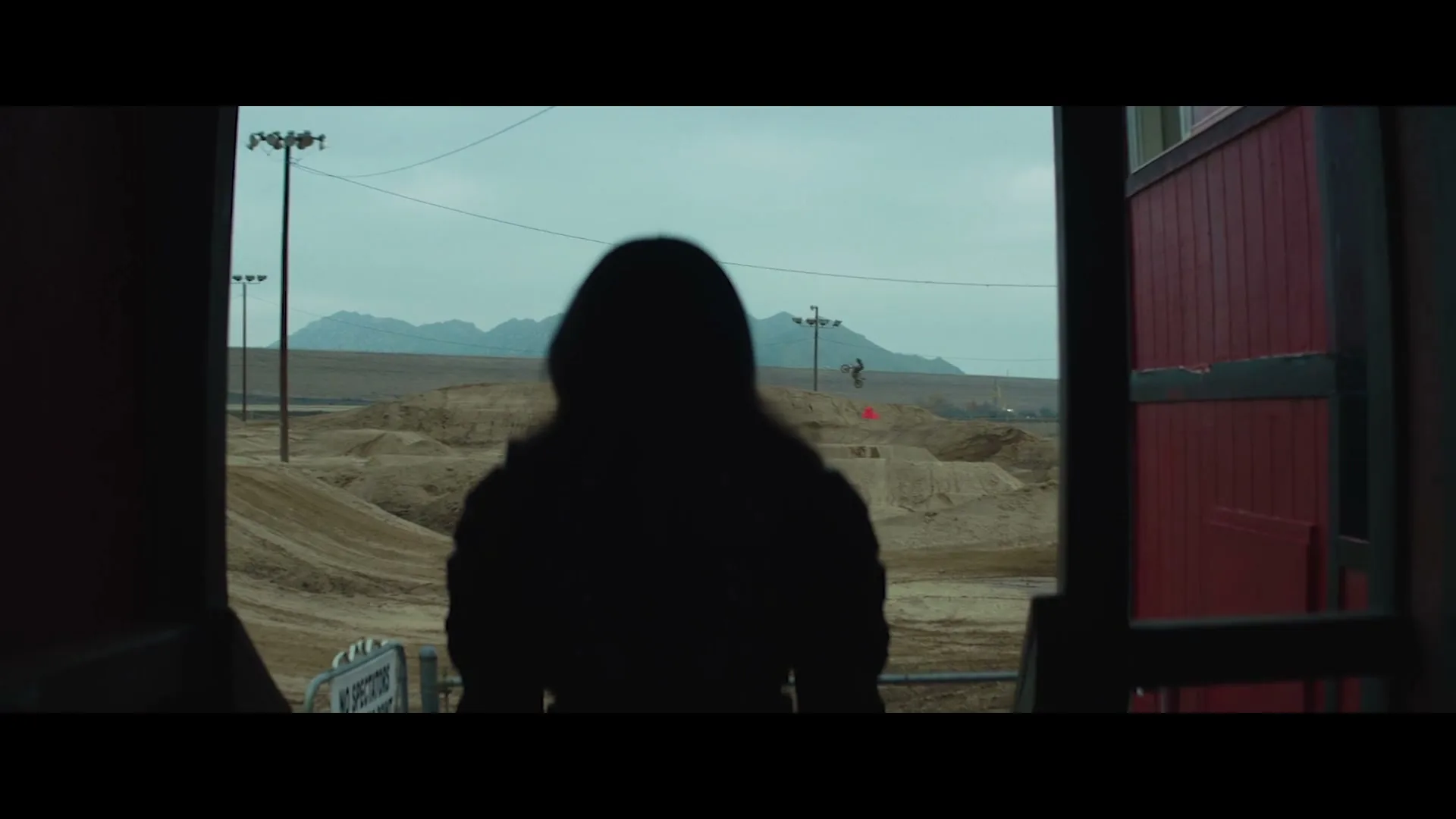A person in silhouette stands in a doorway, looking out at a vast, dusty motocross track with mountains in the distance under a cloudy sky.