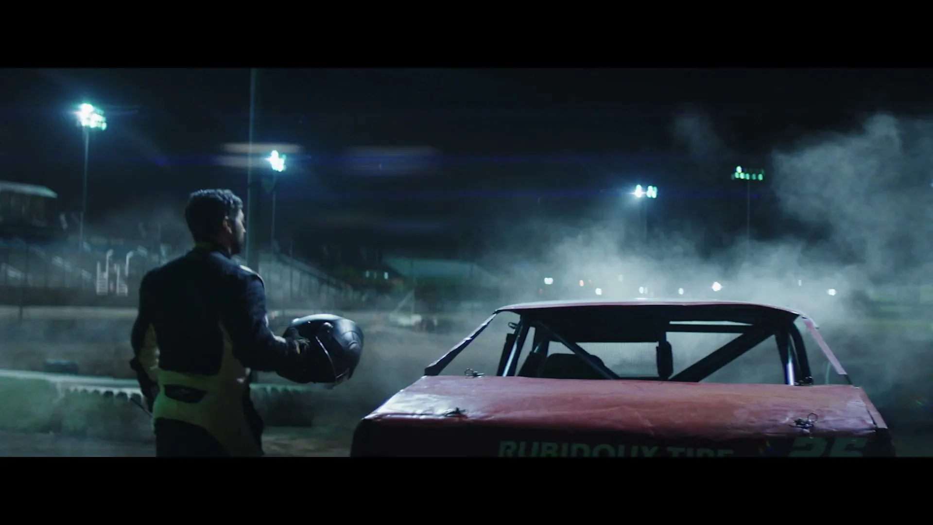 A male race car driver in a racing suit stands next to his red stock car, holding his helmet, at a dirt track at night.