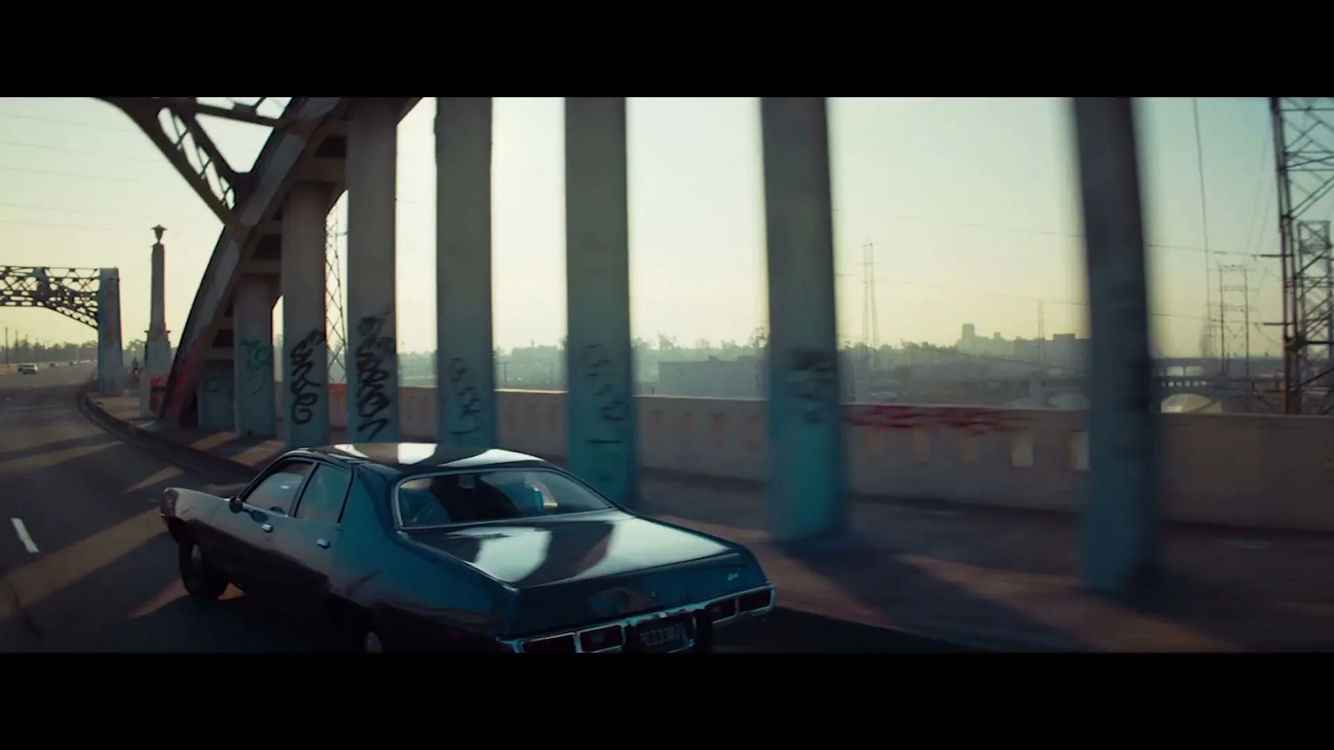A dark vintage car drives across a graffiti-covered bridge at golden hour, with sunlight glinting off its roof and the city skyline in the background.