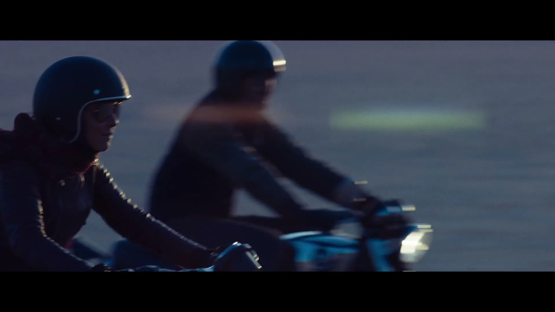 Two people wearing helmets ride motorcycles side-by-side on a road at dusk, with the person in the foreground smiling.