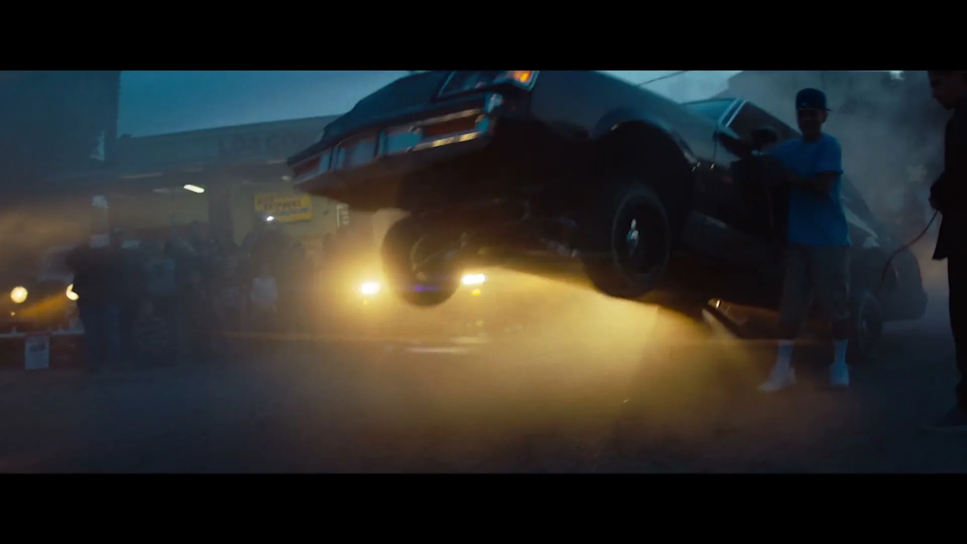 Two men stand next to a dark lowrider car that bounces on its hydraulic suspension, surrounded by smoke and onlookers at dusk.