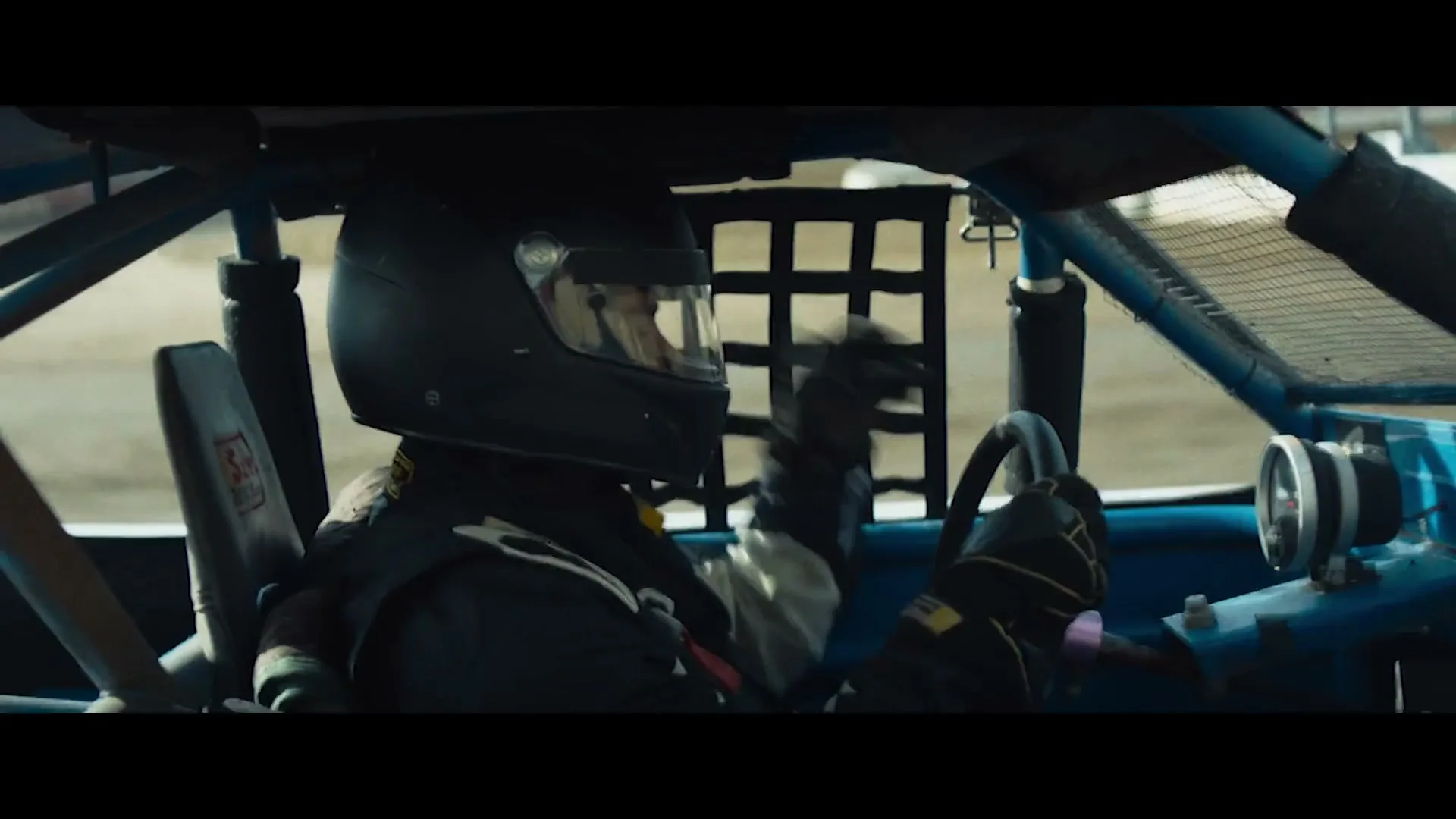 A race car driver, wearing a black helmet and racing suit, is seen from the side, driving a blue race car on a dirt track during the day.
