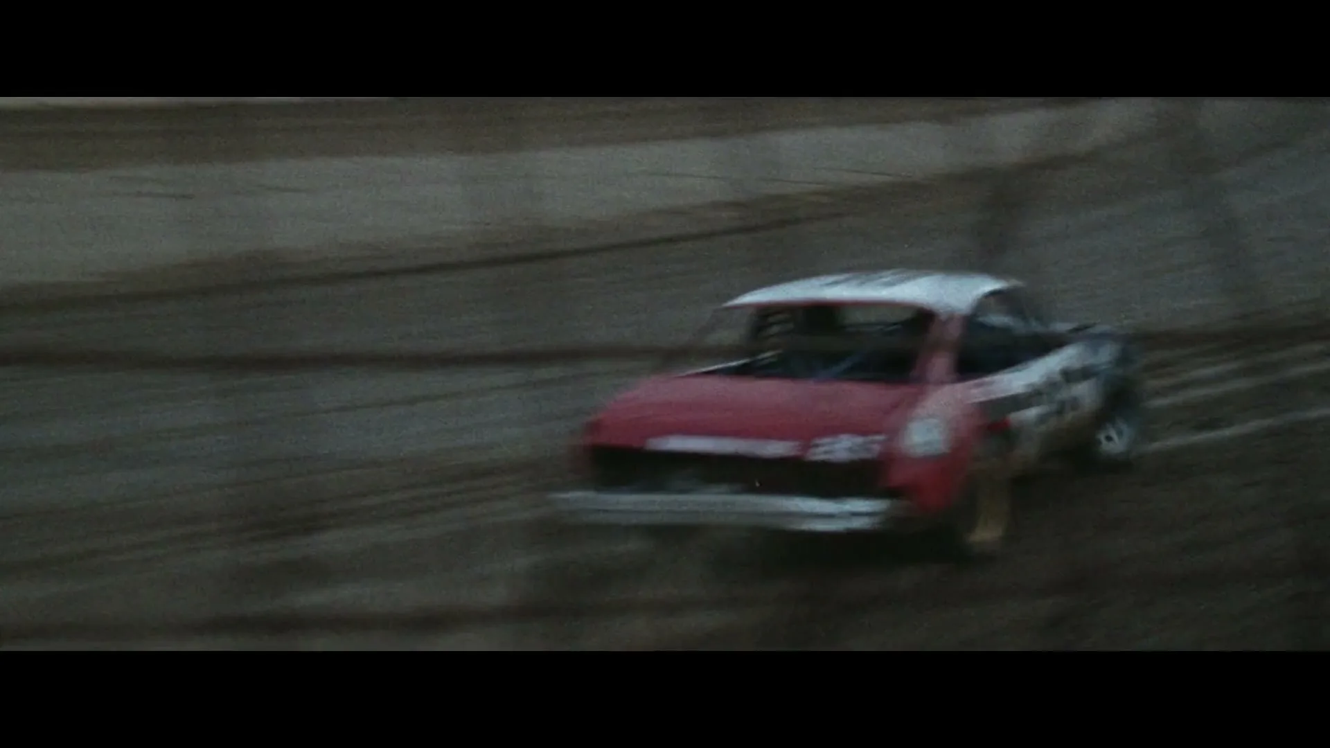 A red and white stock car races on a dirt track, kicking up mud and dust, with other vehicles visible in the distance under a cloudy sky.
