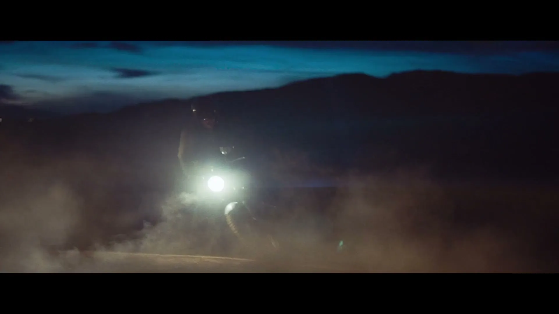 A person on a motorcycle with a bright headlight kicks up dust on a dark, mountainous road at night.