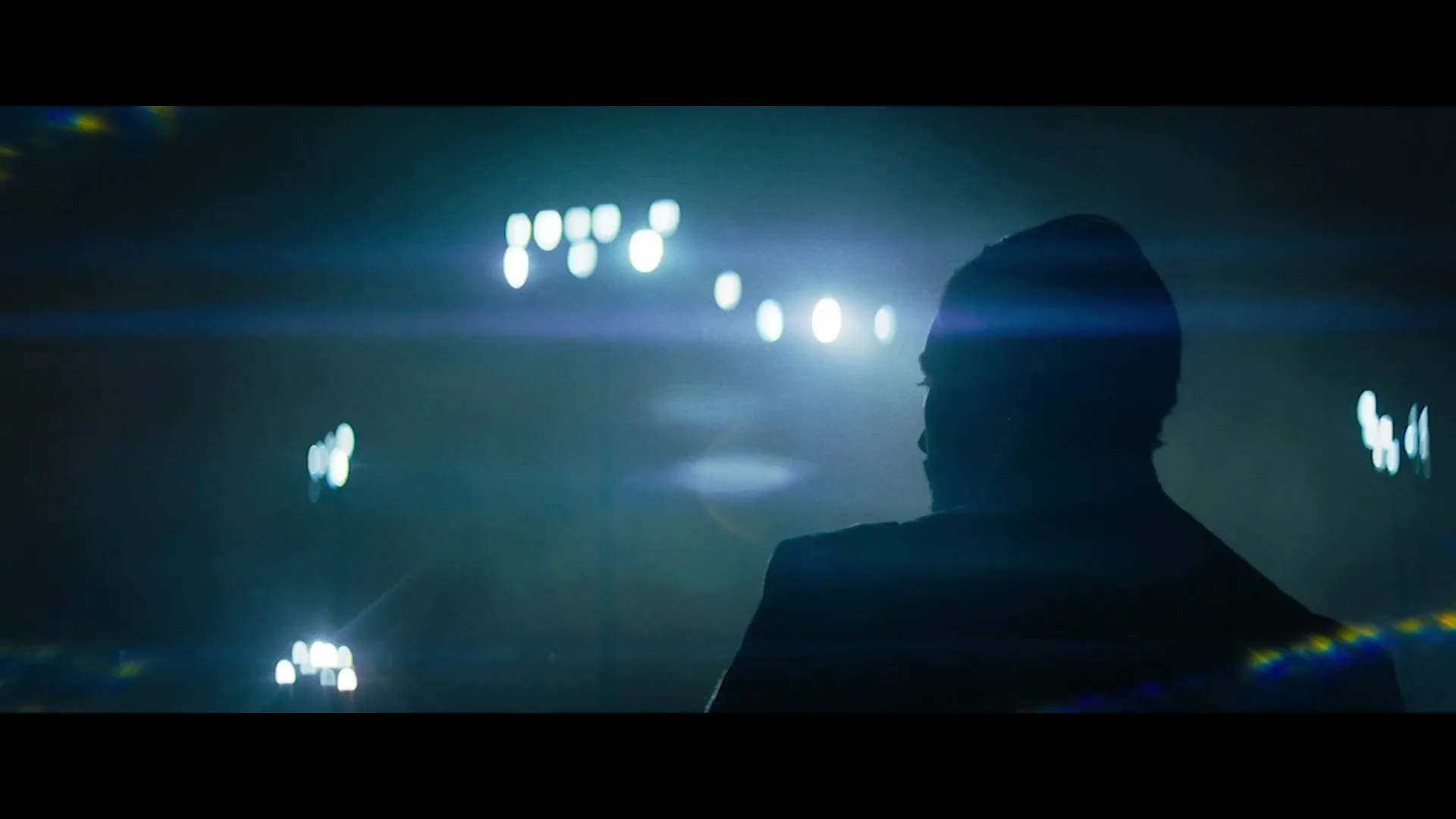 A silhouetted man with a beard and beanie looks out at bright stadium lights in a dark, hazy arena.