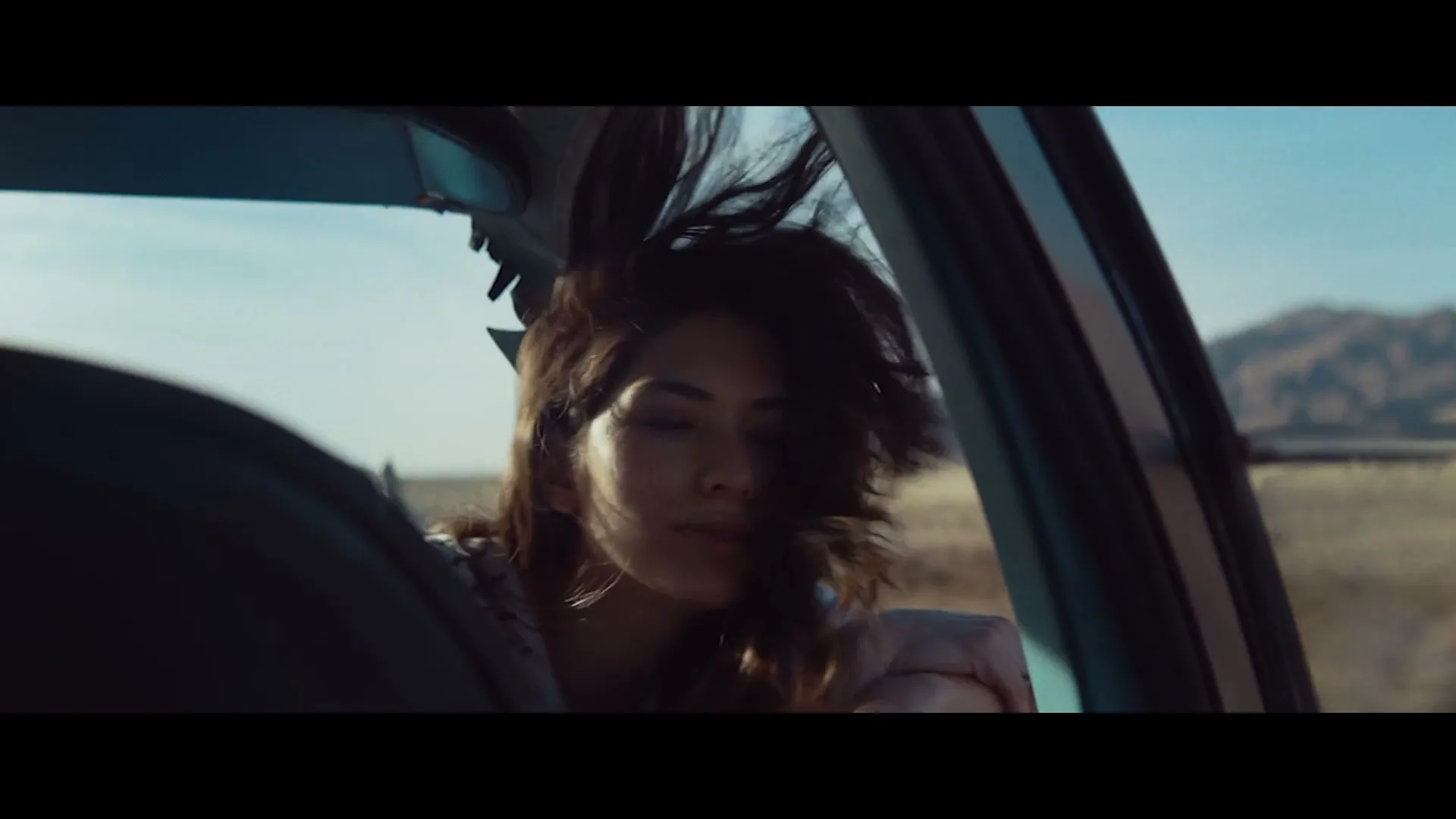 A young woman with dark hair leans out of a car window, her hair blowing in the wind as she drives through a desert landscape.