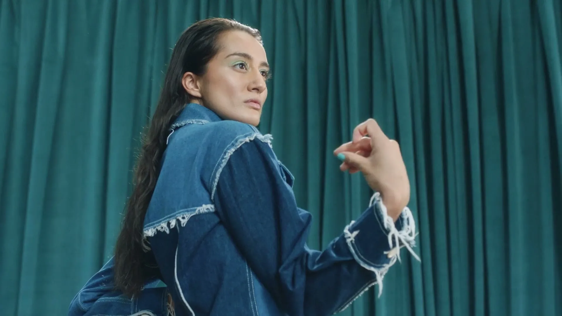 A woman with long dark hair and a denim jacket looks away from the camera, gesturing with her hand against a teal curtain background.