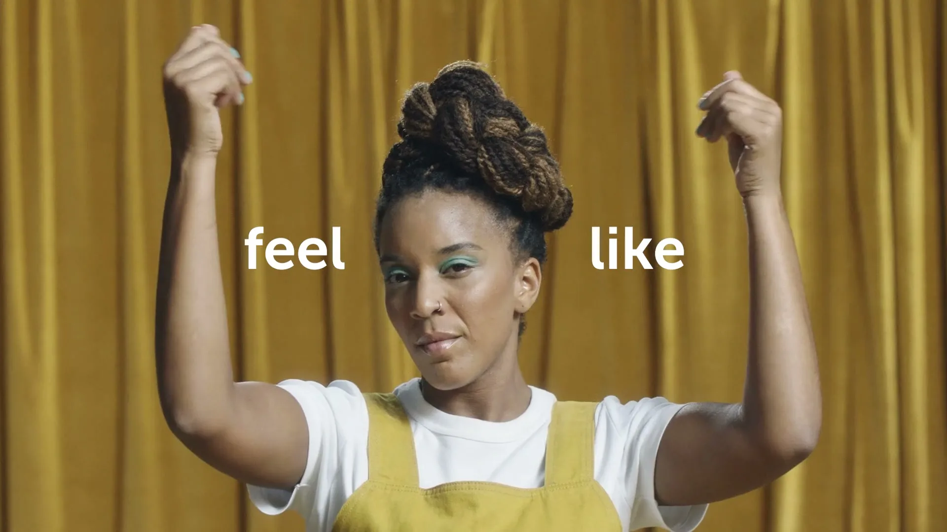 A woman with braided hair in a bun, wearing a white t-shirt and yellow overalls, raises her arms in front of a yellow curtain.