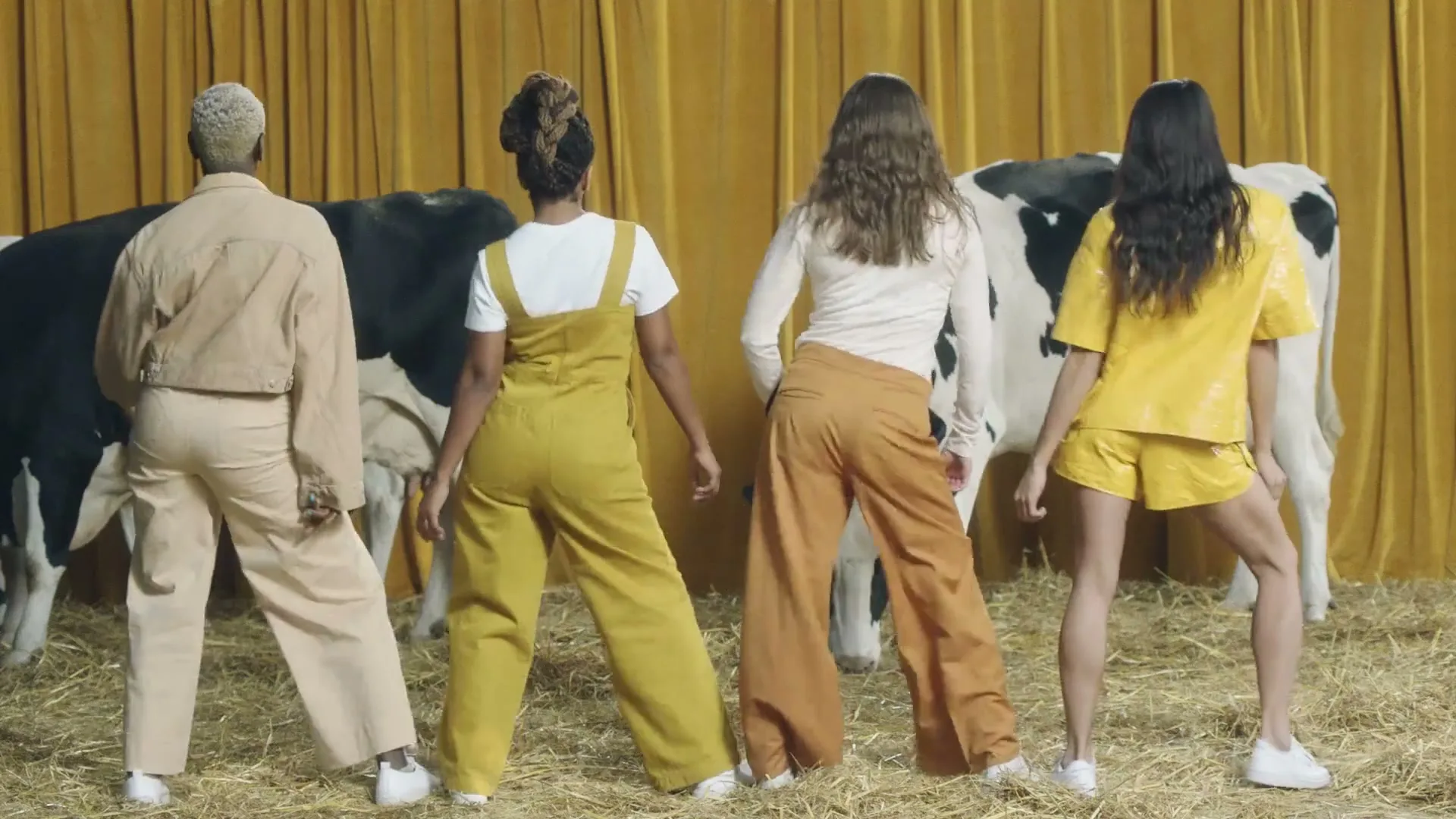 Four women in earth-toned outfits dance with their backs to the camera, in a barn with two cows and a golden curtain.