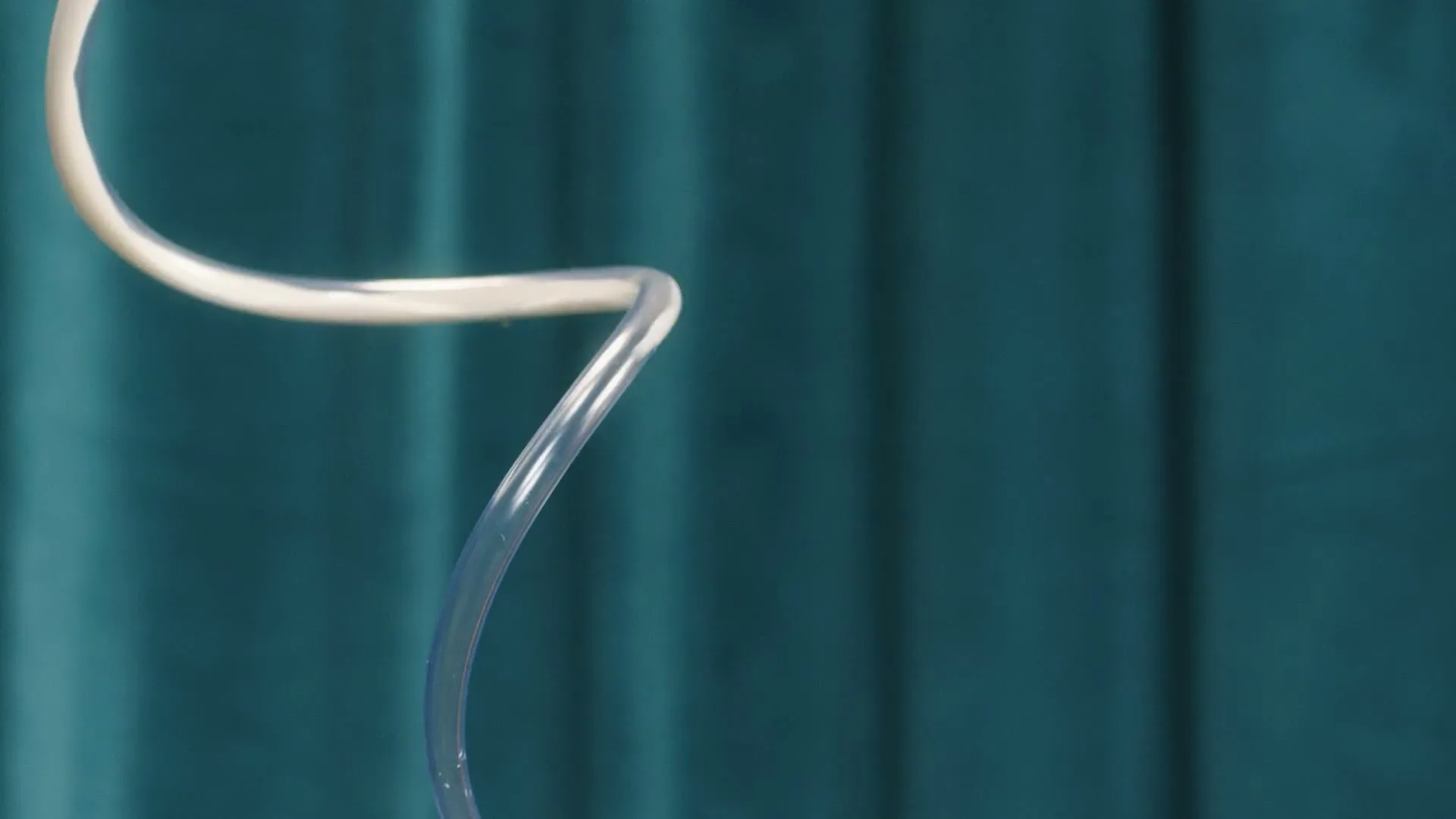A clear plastic tube, possibly for medical use, is shown against a teal curtain, with a white tube appearing to replace it.