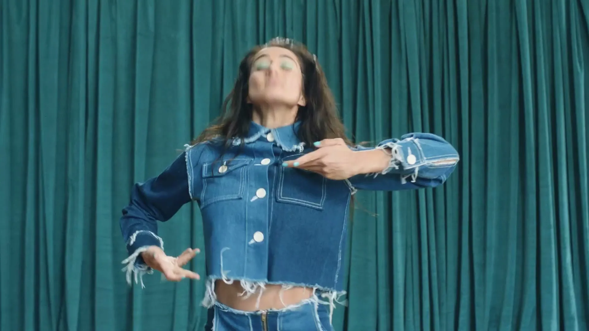 A woman with long dark hair and a denim outfit dances energetically in front of a teal curtain, moving her arms and body.