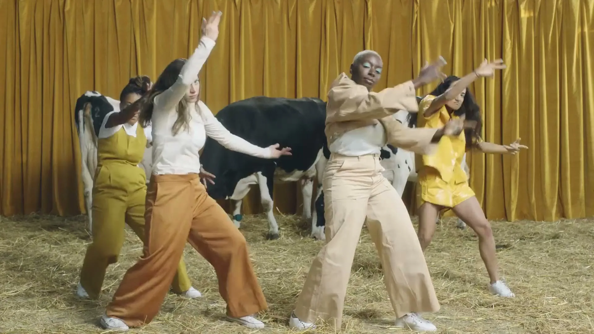 Four diverse women in casual attire dance energetically in a barn setting with cows and a golden curtain backdrop.