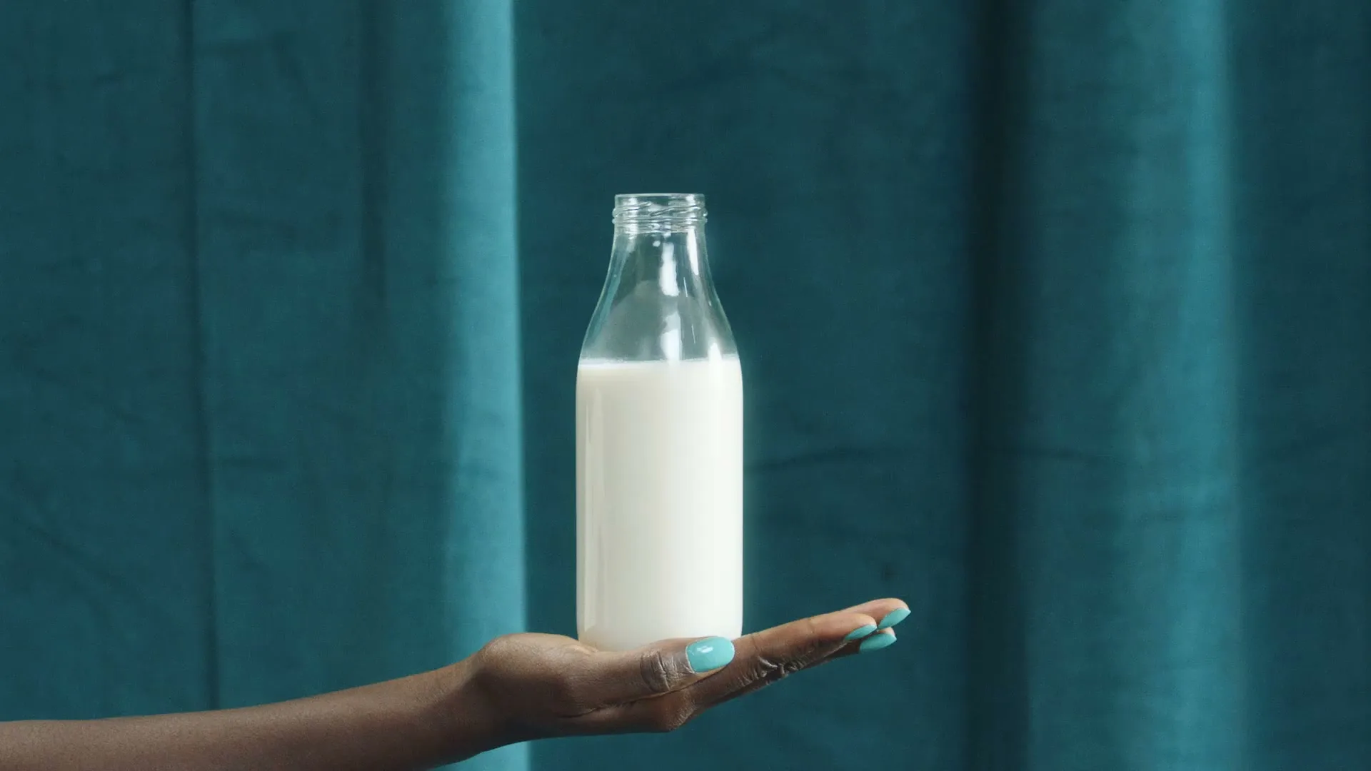 A dark-skinned hand with teal nail polish holds an empty glass milk bottle that magically fills with milk against a teal curtain background.