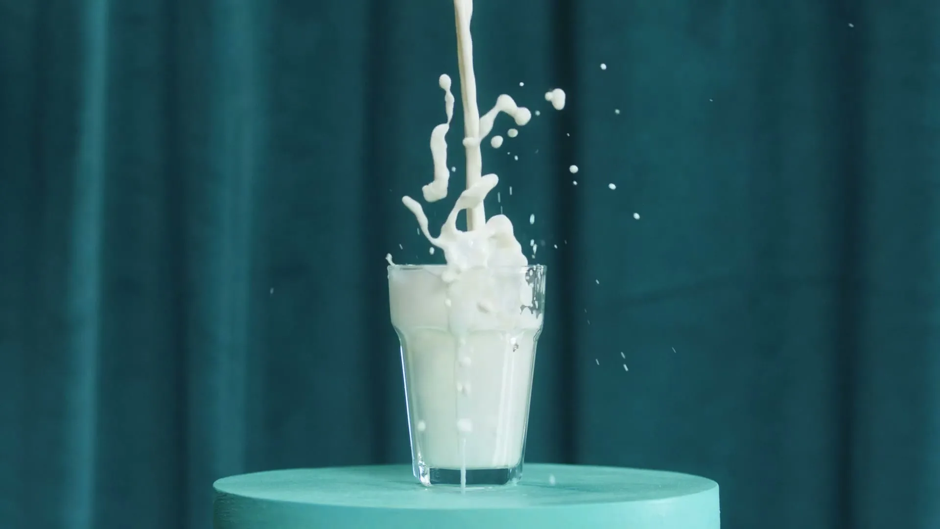 Milk is poured into a glass, overflowing and splashing onto a teal pedestal against a teal curtain.