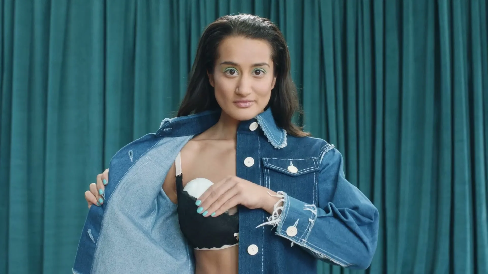 A woman in her 20s, wearing a denim jacket and a black bra, reveals a breast pump and then holds it up to her face, in a studio.