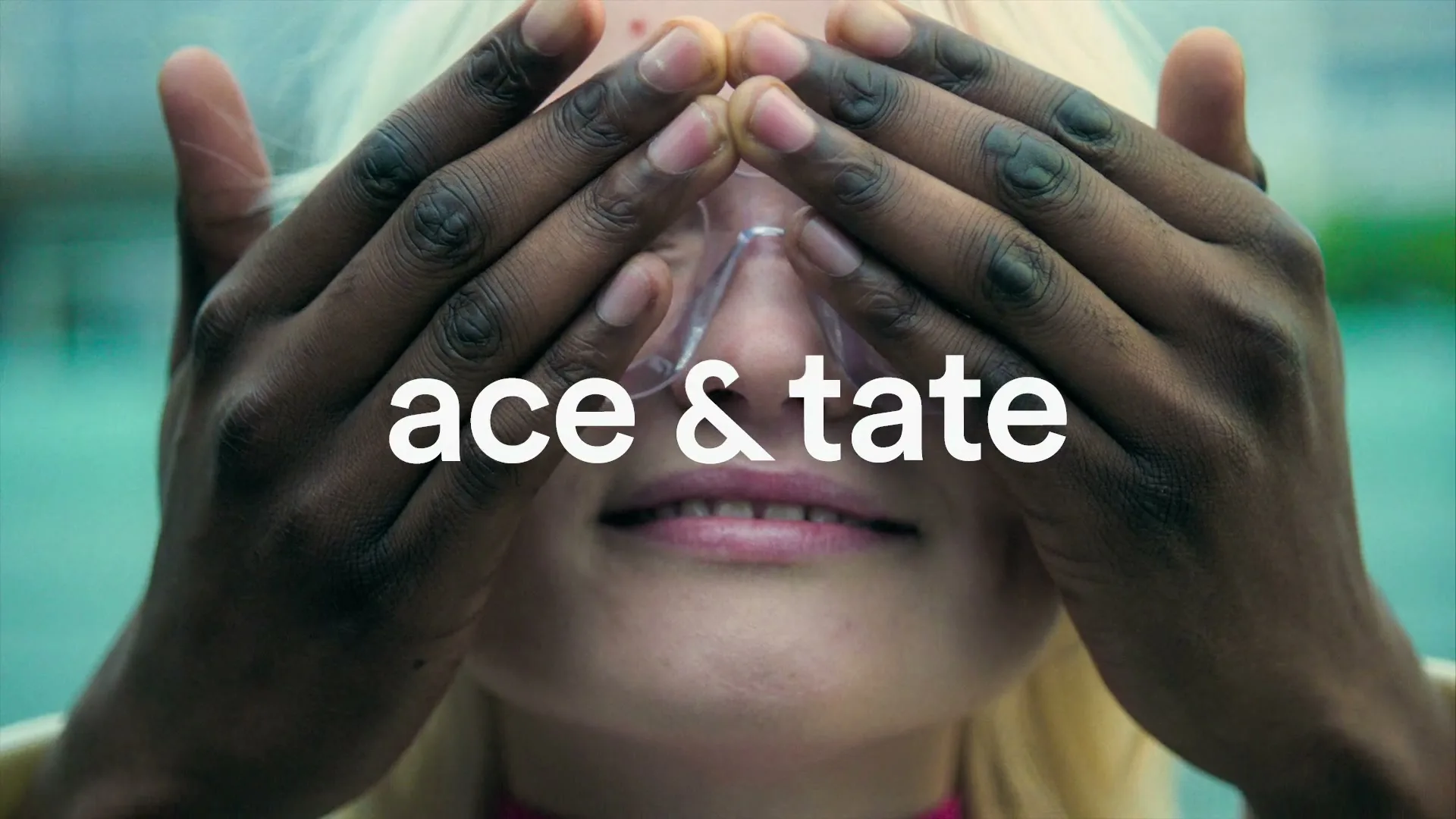 A blonde woman with clear glasses smiles as dark-skinned hands remove from her eyes, with the brand logo overlaid.