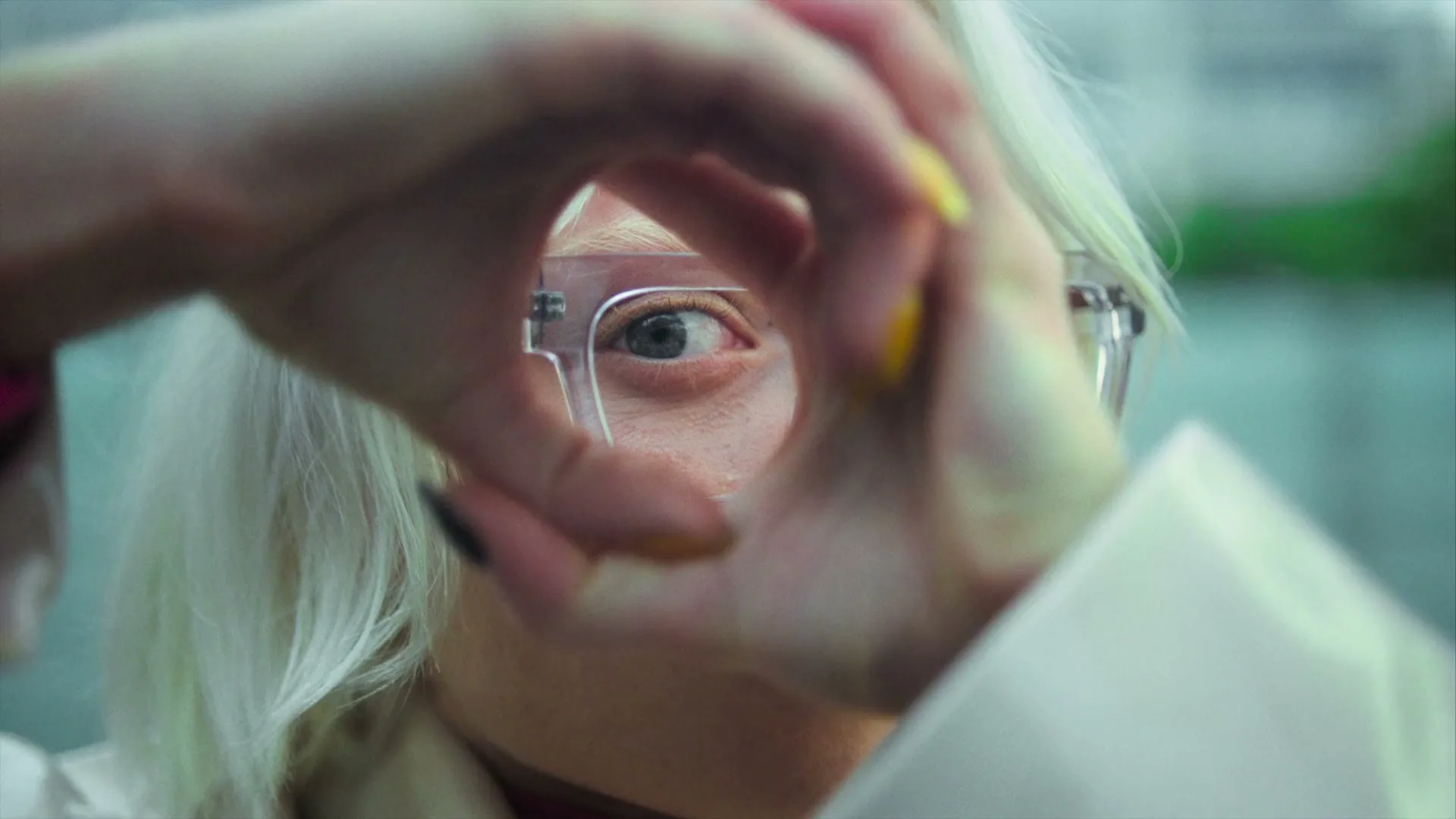 A person with white hair and glasses looks through a finger circle, with their eye visible through the lens.