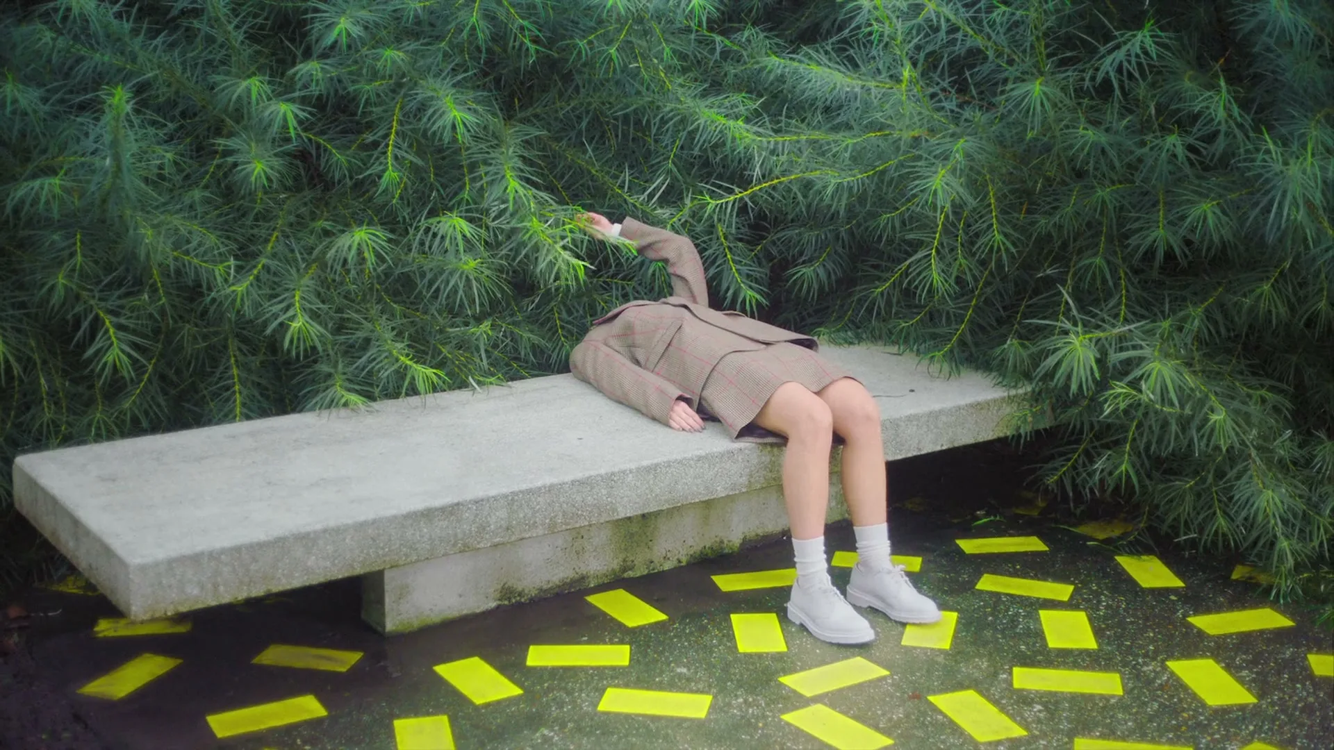 A person in a brown suit and skirt, with no head, lies on a concrete bench, surrounded by green foliage and yellow rectangles on the ground.