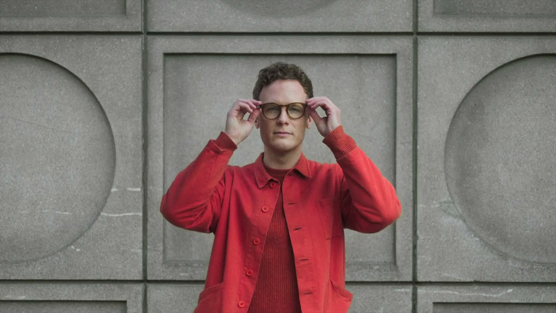 A man in a red shirt and jacket adjusts his glasses while looking directly at the camera, standing against a concrete wall with geometric patterns.