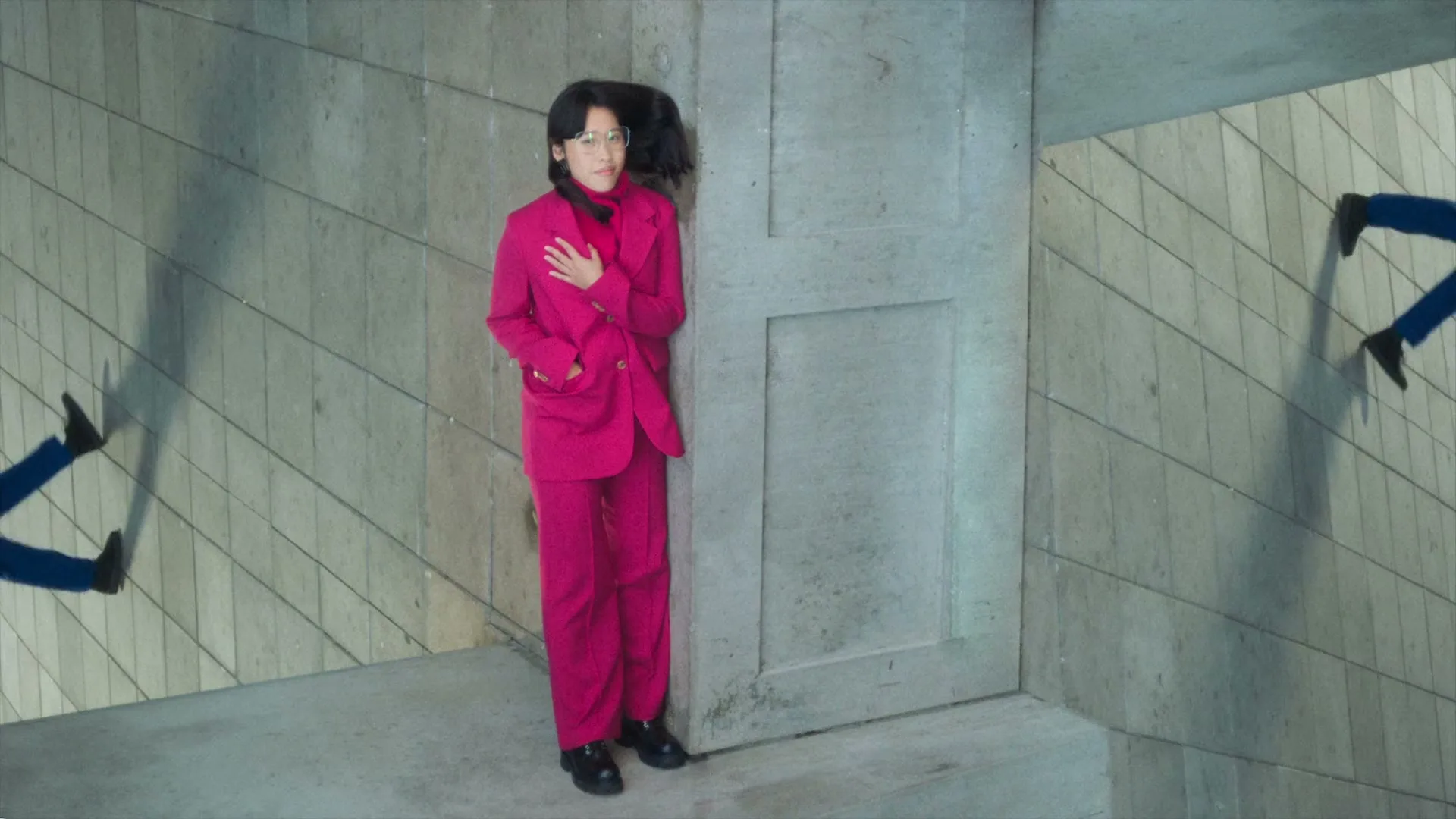 A person in a bright pink suit and glasses stands in a corner, with their hand on their chest, as animated legs walk on the walls around them.