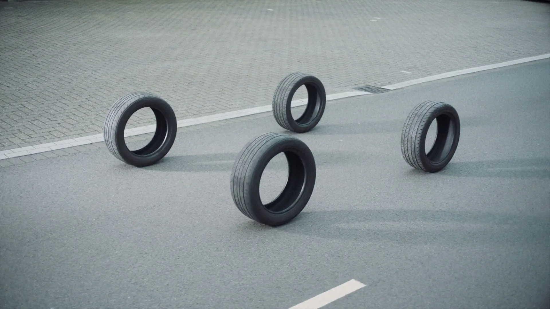 Four car tires are arranged on a grey asphalt road with white markings, creating an abstract and minimalist composition.