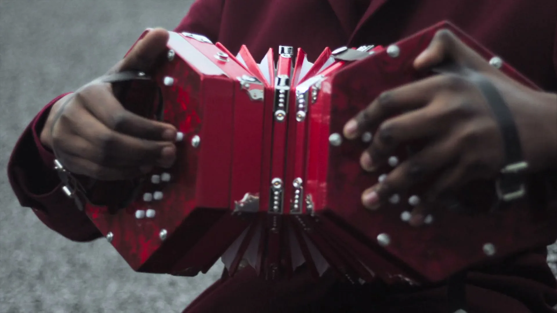 A person with dark skin and a maroon jacket plays a red concertina, with their hands visible on the instrument.