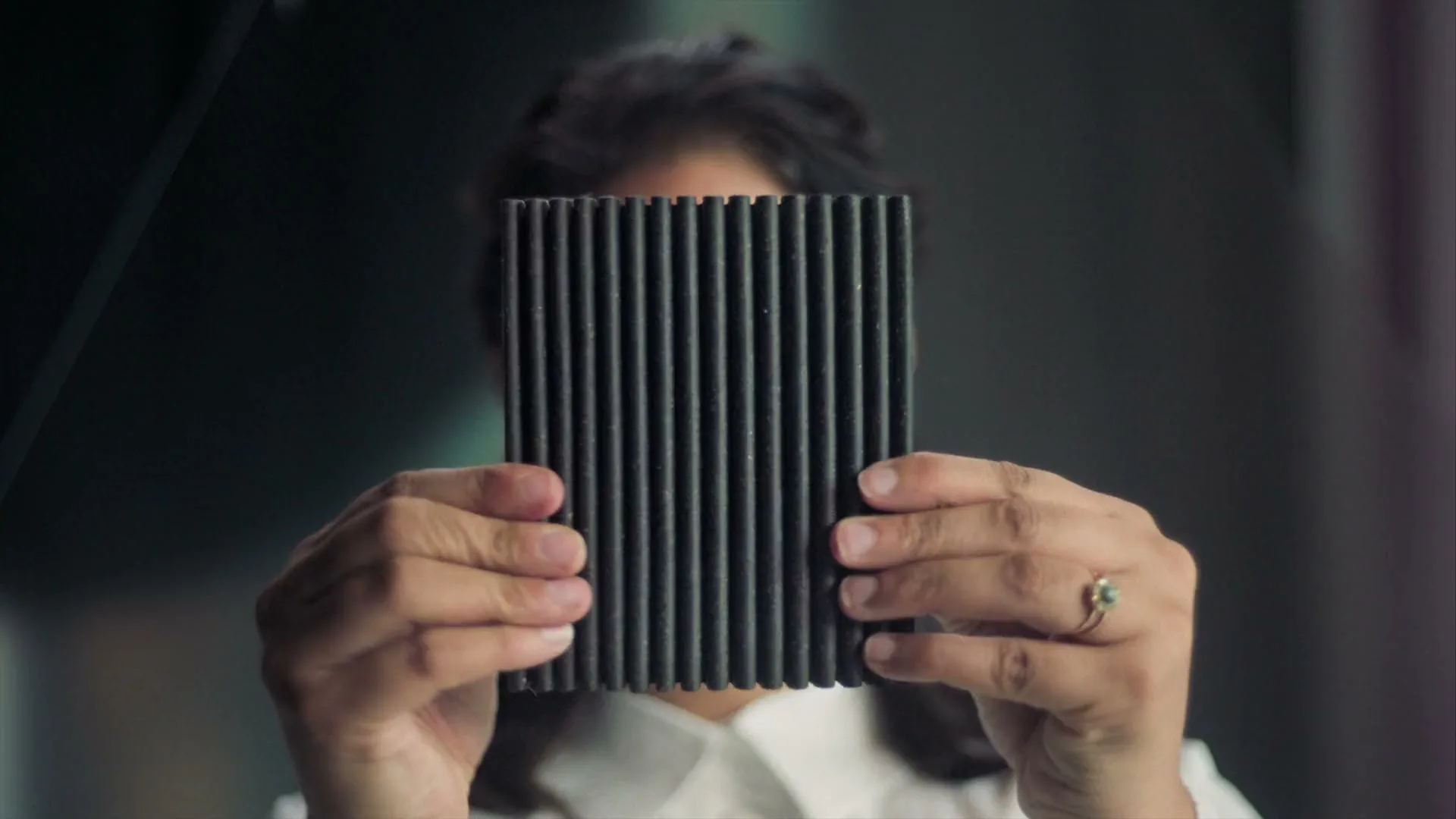 A person with dark hair holds a black corrugated object in front of their face, obscuring it, in an interior setting.