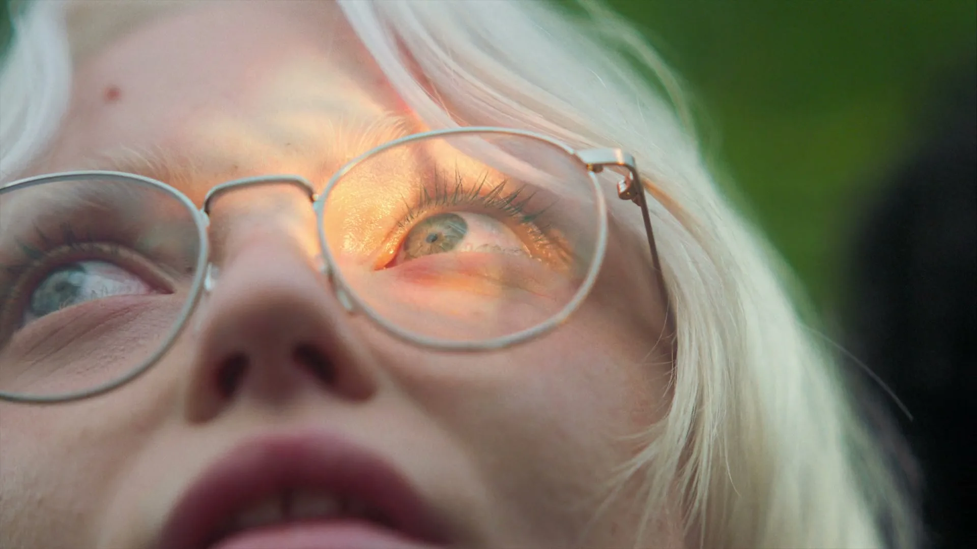 A young woman with platinum blonde hair and round glasses looks up, with sunlight reflecting in her lenses, in an outdoor setting.
