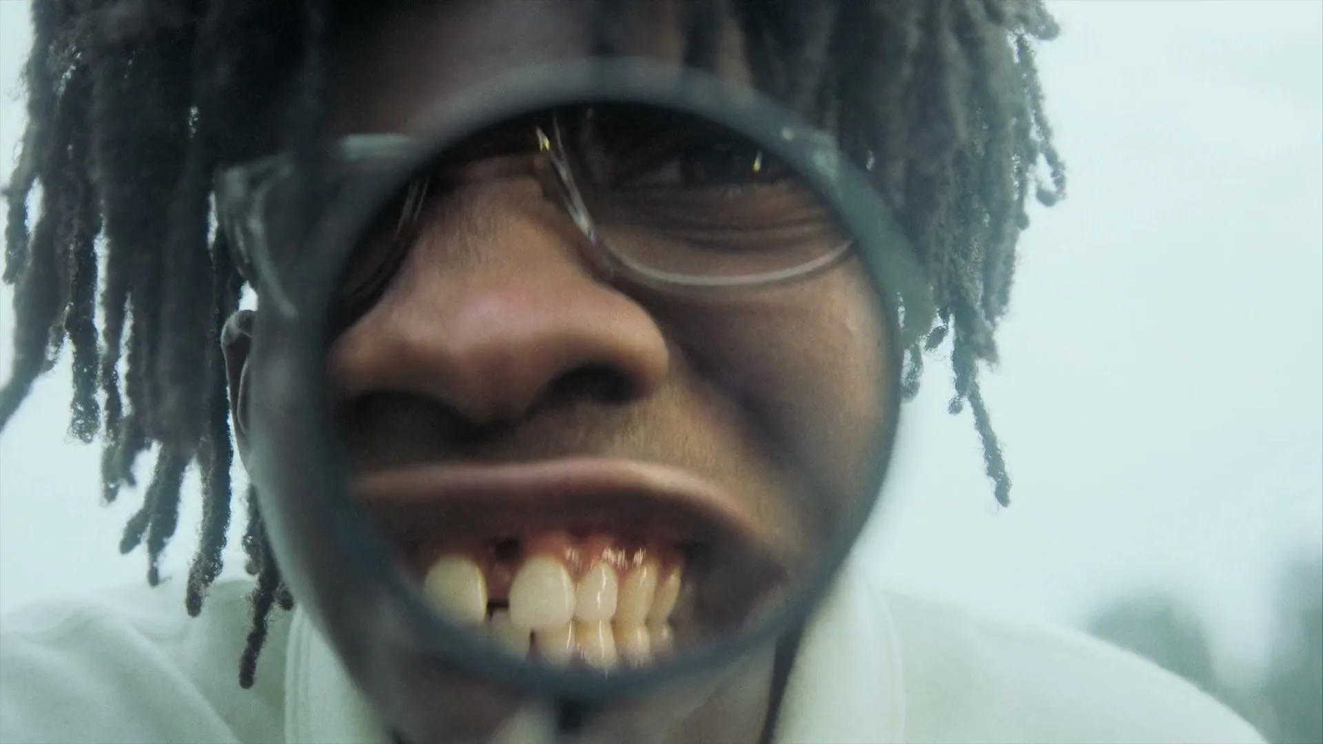 A person with dreadlocks and glasses looks through a magnifying glass, distorting their face, in a bright, hazy outdoor setting.
