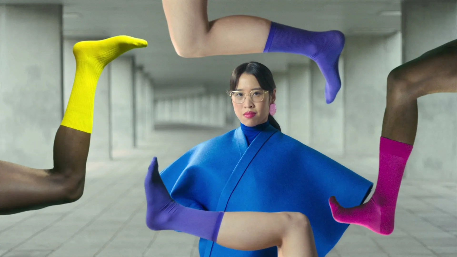 A woman in a blue outfit and glasses looks at the camera, surrounded by disembodied legs wearing colorful socks in a concrete hallway.