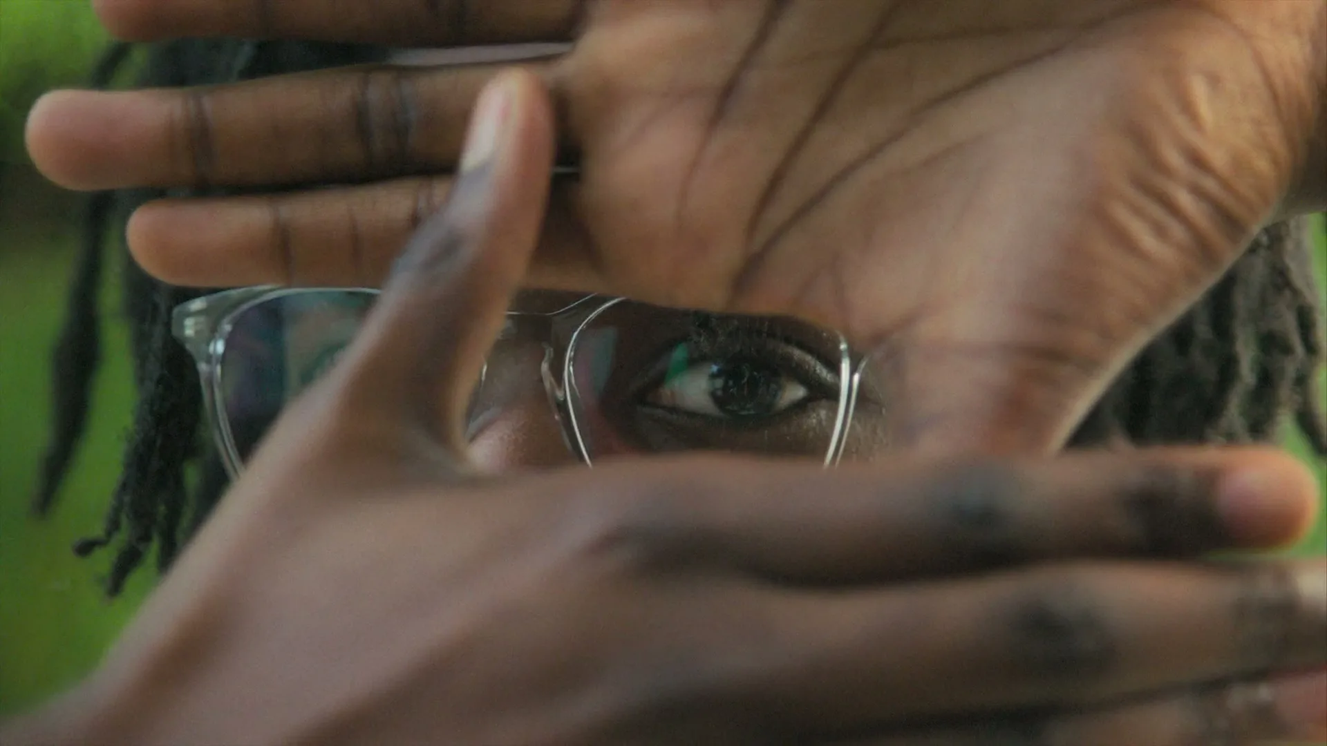 A person with dreadlocks and clear glasses frames their eye with their hands, looking directly at the camera against a green background.