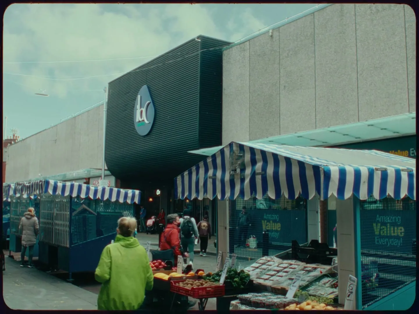 A wide shot of a bustling outdoor market in front of a shopping center, with people walking around and browsing produce stalls under striped awnings.