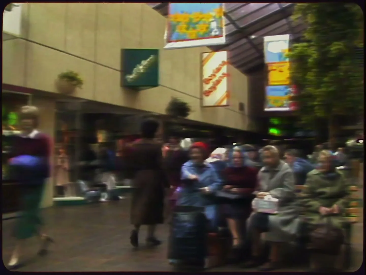 A bustling indoor shopping center with people walking and sitting, featuring shops and hanging banners, captured in a vintage style.