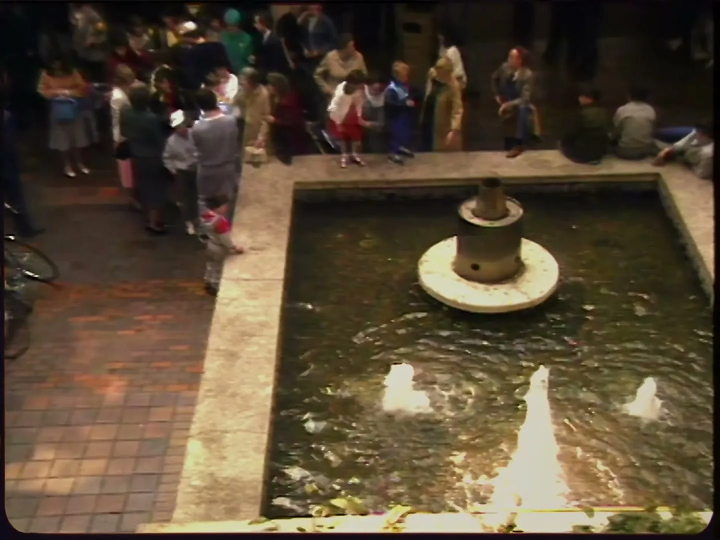 A high-angle shot captures a bustling public square with a central fountain, surrounded by diverse people of all ages, some standing, some sitting.