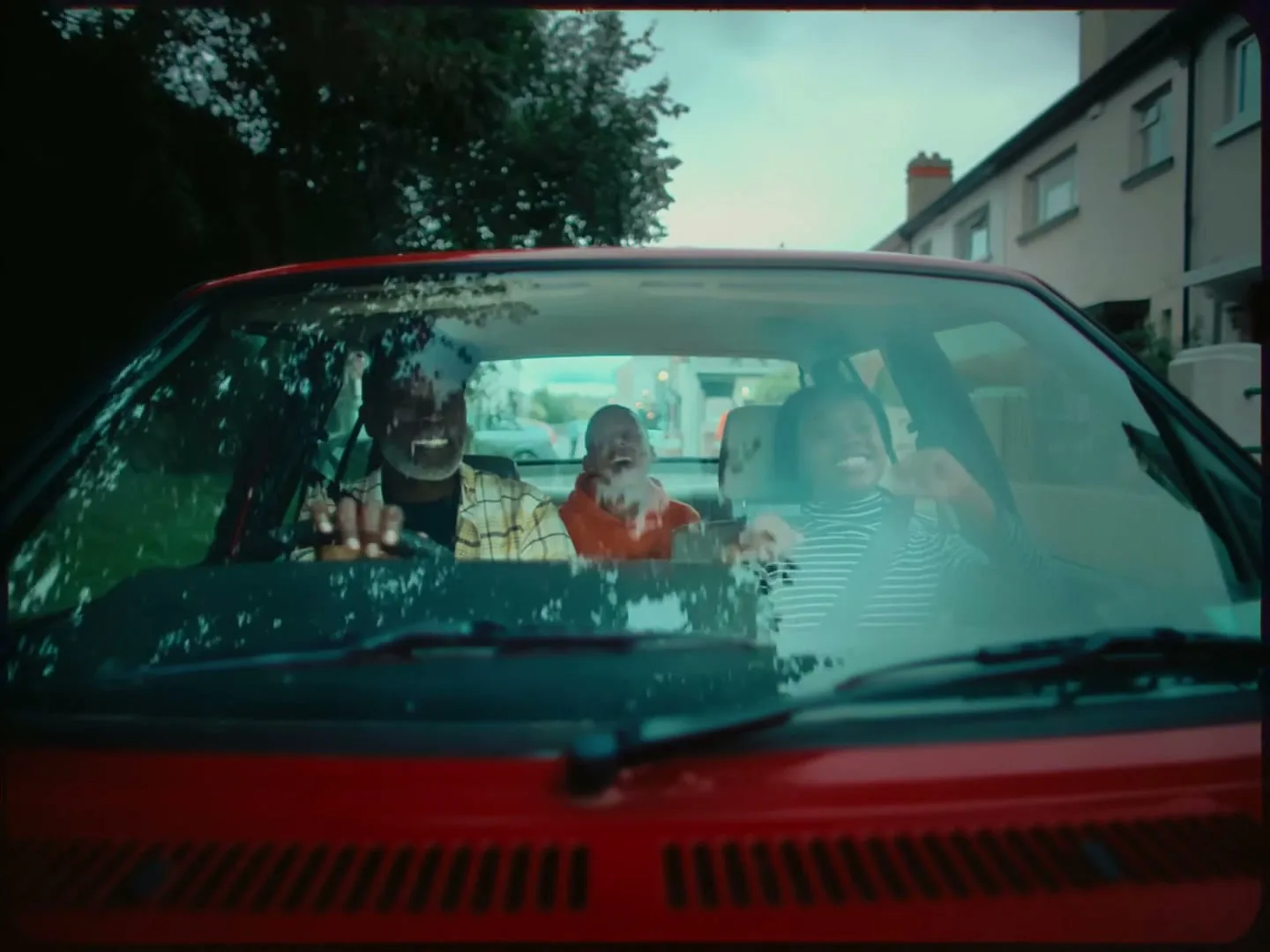 A family of three, a man, a woman, and a child, are seen through the windshield of a red car, smiling and laughing as they drive down a residential street.