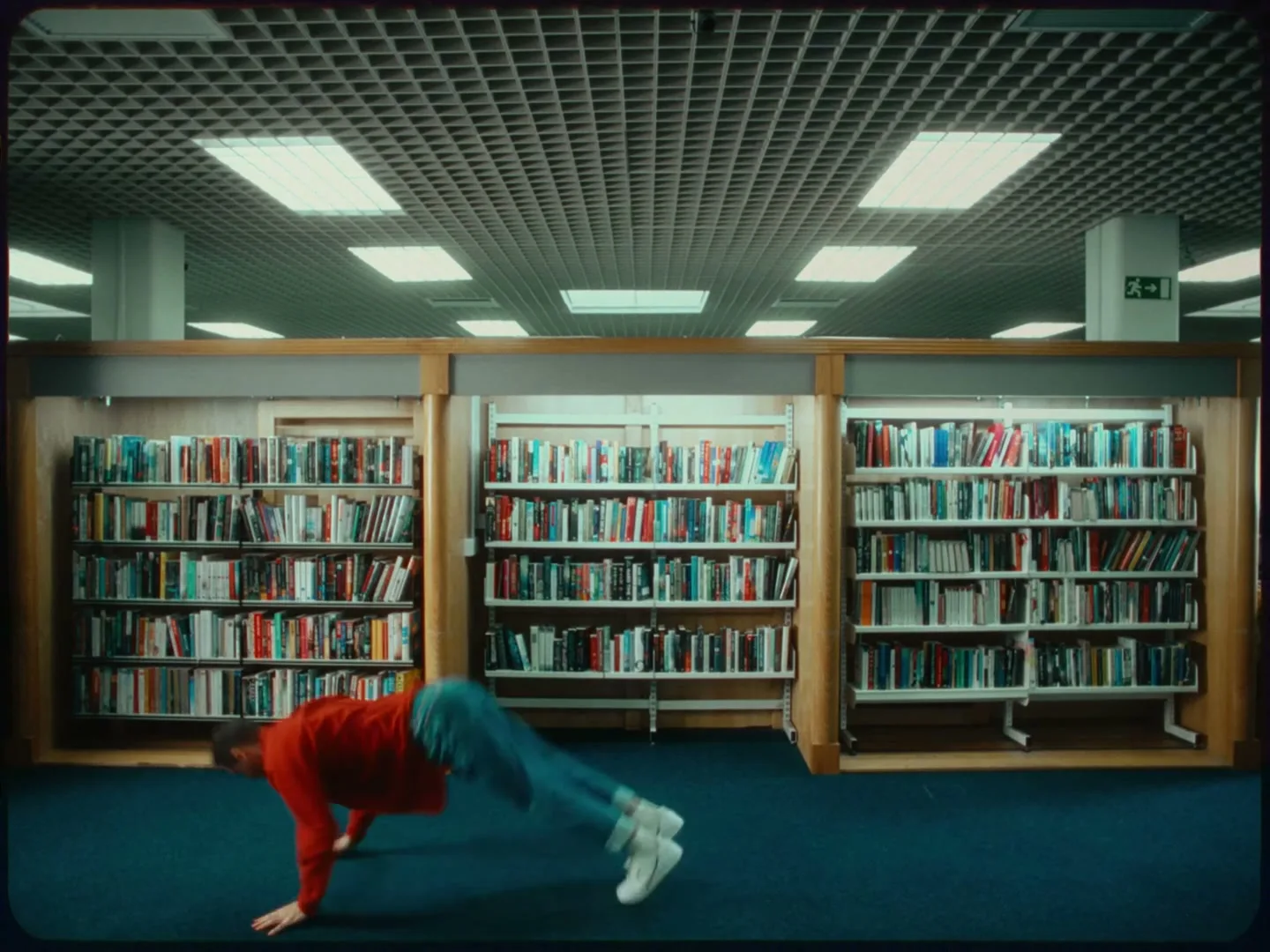 A young man in a red top and blue jeans breakdances on a blue carpeted floor in a library, surrounded by bookshelves.