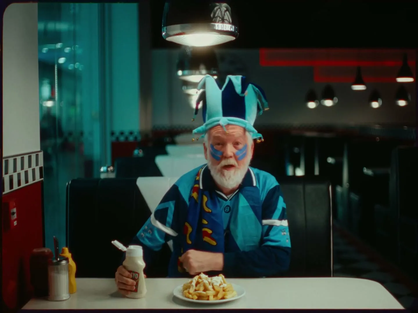 A senior man with a white beard and a jester hat squeezes mayonnaise onto a plate of french fries in a diner booth.