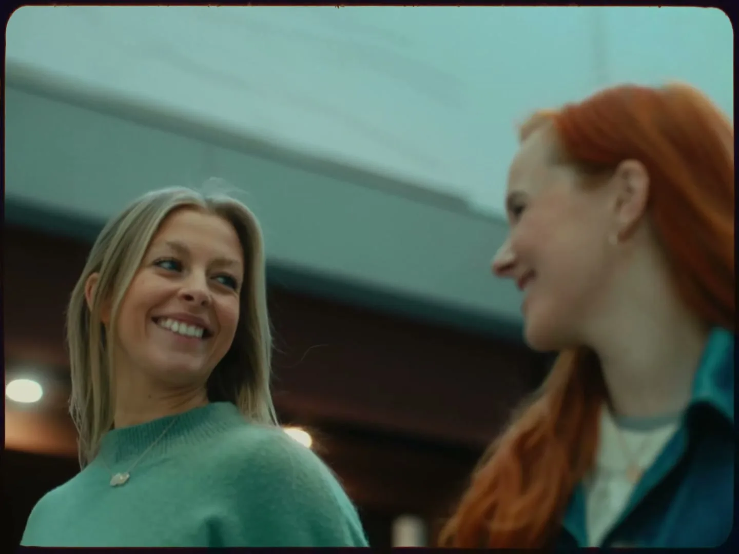 Two women, one with blonde hair and a green sweater, the other with red hair, smile and laugh together in an indoor public space.