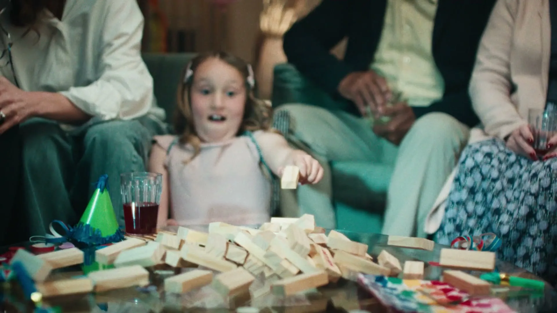 A young girl in a pink dress plays Jenga with adults on a coffee table, looking surprised as the tower collapses during a party.