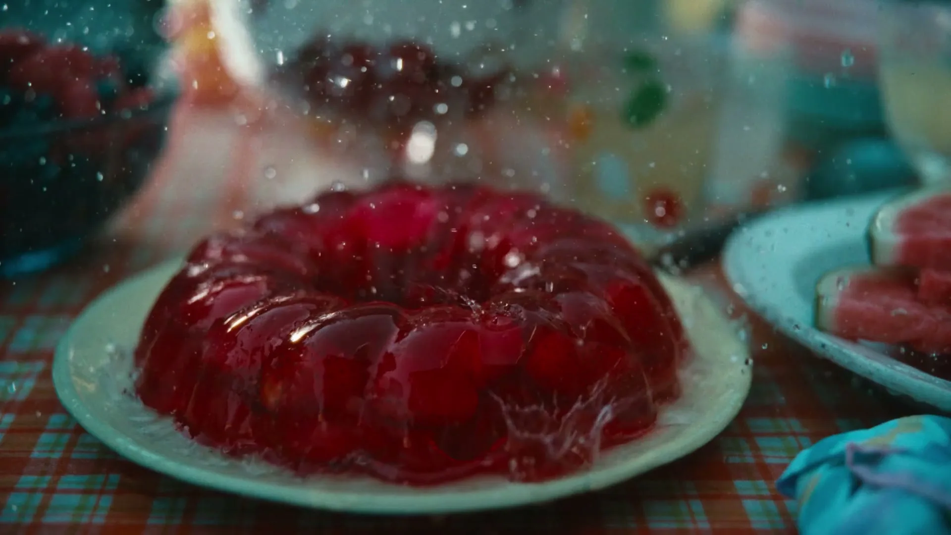 A vibrant red jelly mold sits on a white plate on a checkered tablecloth, with water droplets splashing around it, suggesting a lively outdoor meal.