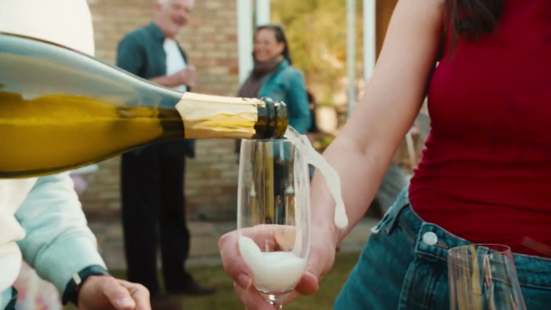 A person in a red top and denim shorts pours champagne into a glass, with other people socializing in the blurred background at an outdoor gathering.