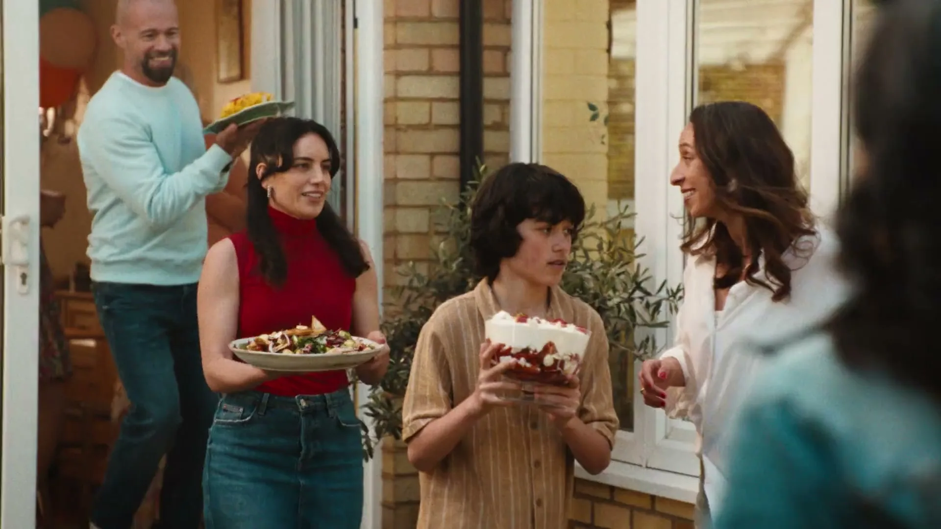 A diverse group of people, including a bald man, a woman in a red top, and a boy, carry food dishes out of a house into a sunny garden party.