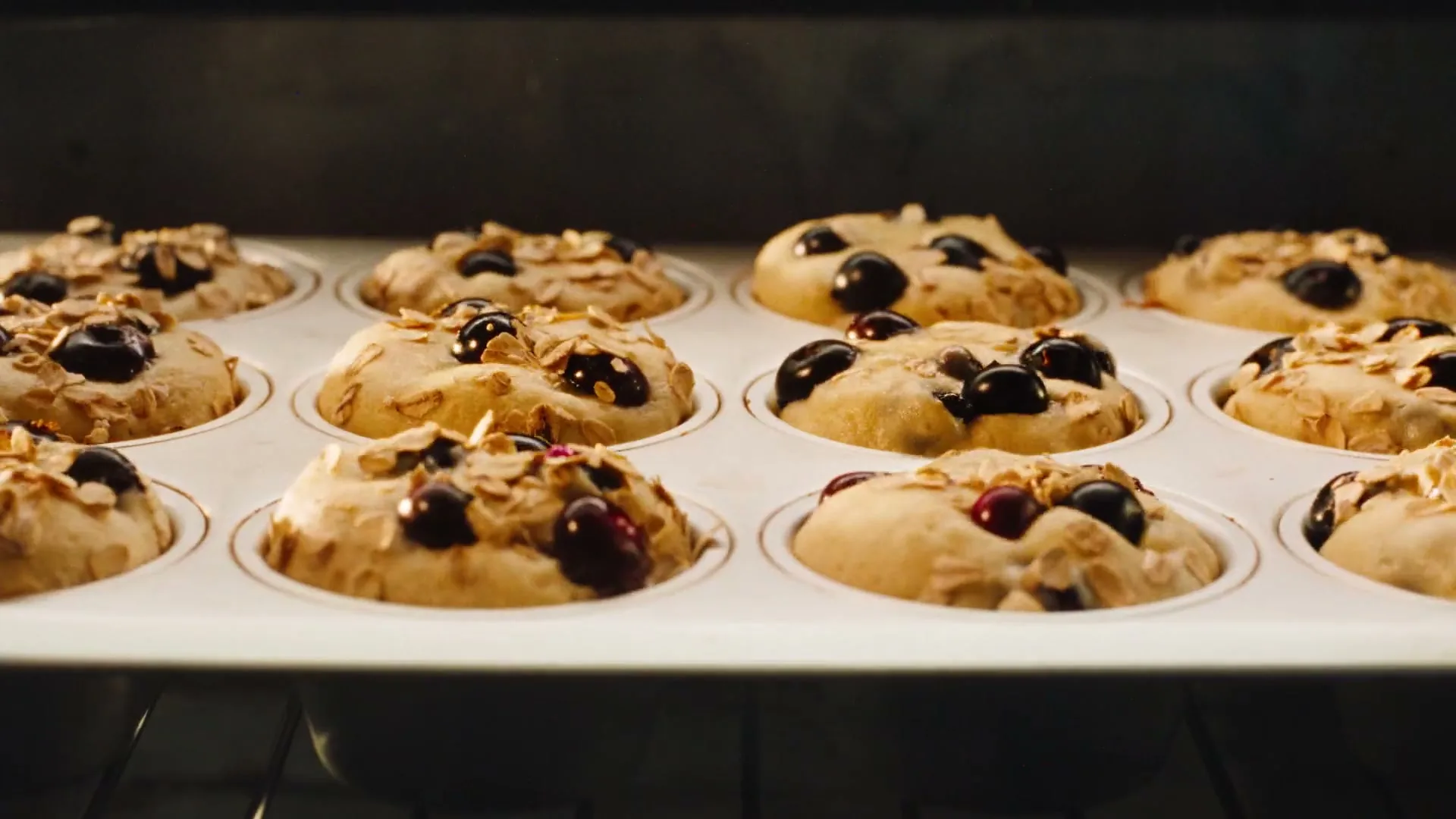 A close-up shot shows a tray of blueberry muffins baking in an oven, with the batter rising and berries visible.