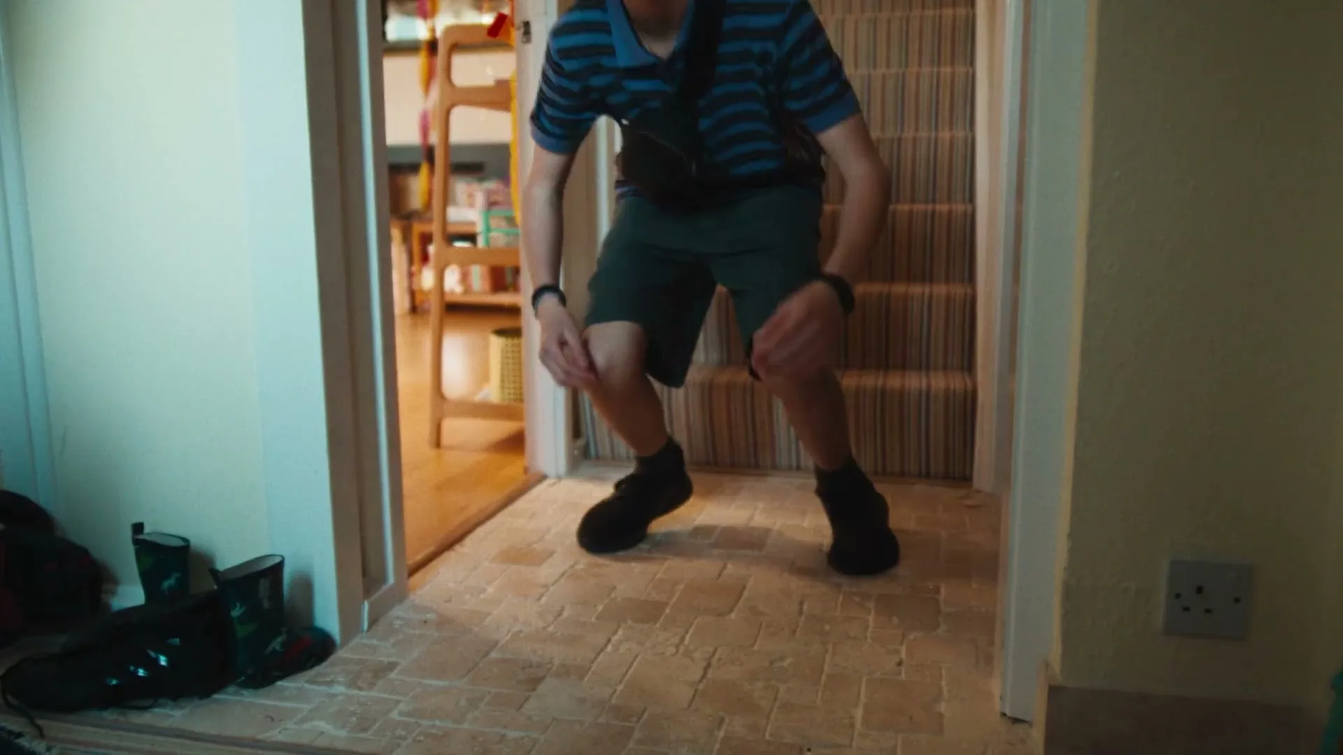 A young male in a striped shirt and shorts jumps excitedly in a hallway with a staircase, celebrating.