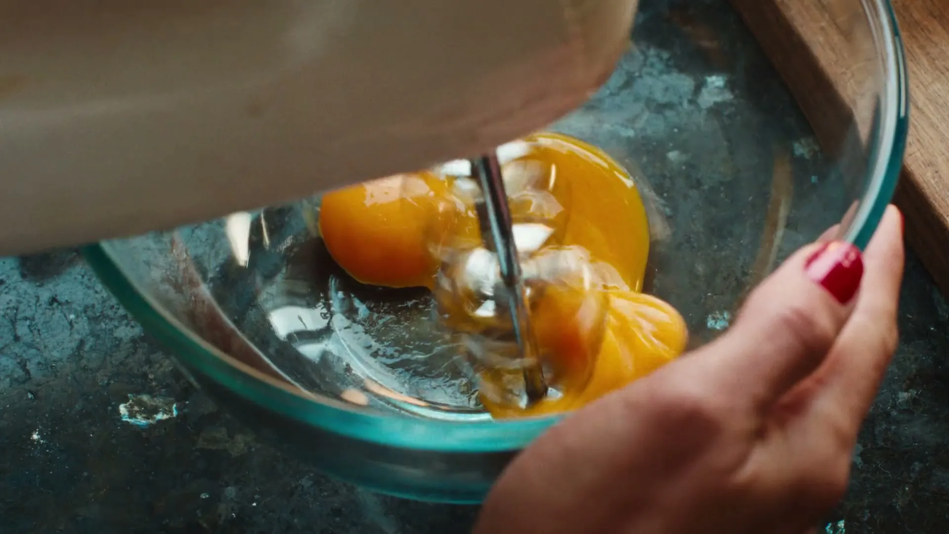 A woman's hand with red nail polish holds a glass bowl as an electric mixer separates three egg yolks from the whites.